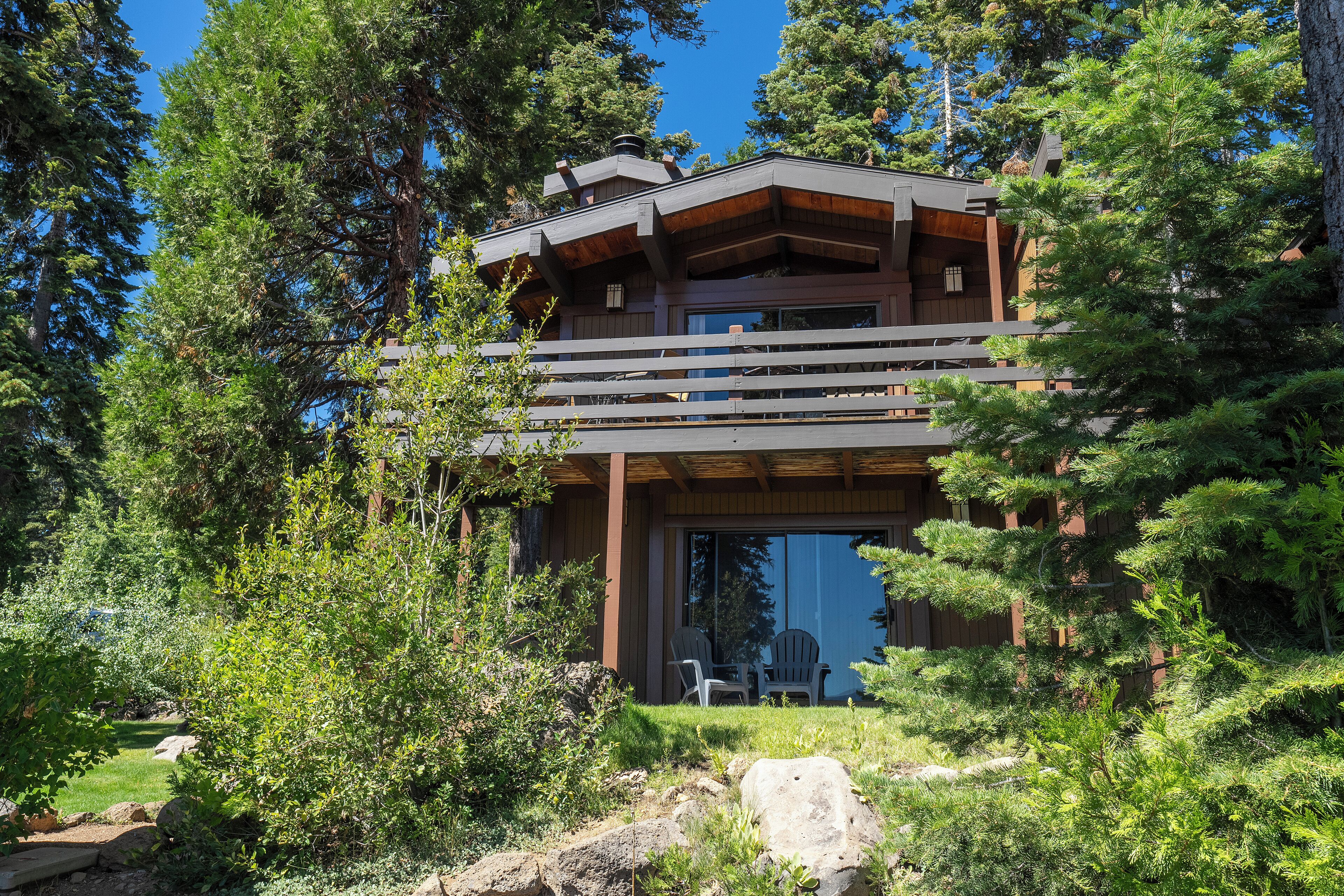 A two-story wooden cabin with a balcony, surrounded by dense forest.