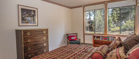 A bedroom with a large bed, a dresser, a chair, a bookshelf, and a window with a view of trees.