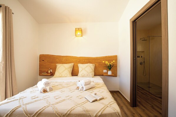 Una habitación de hotel con cabecera de madera, cama con colcha color beige, mesita de noche con un jarrón de flores y ducha con mampara de vidrio.