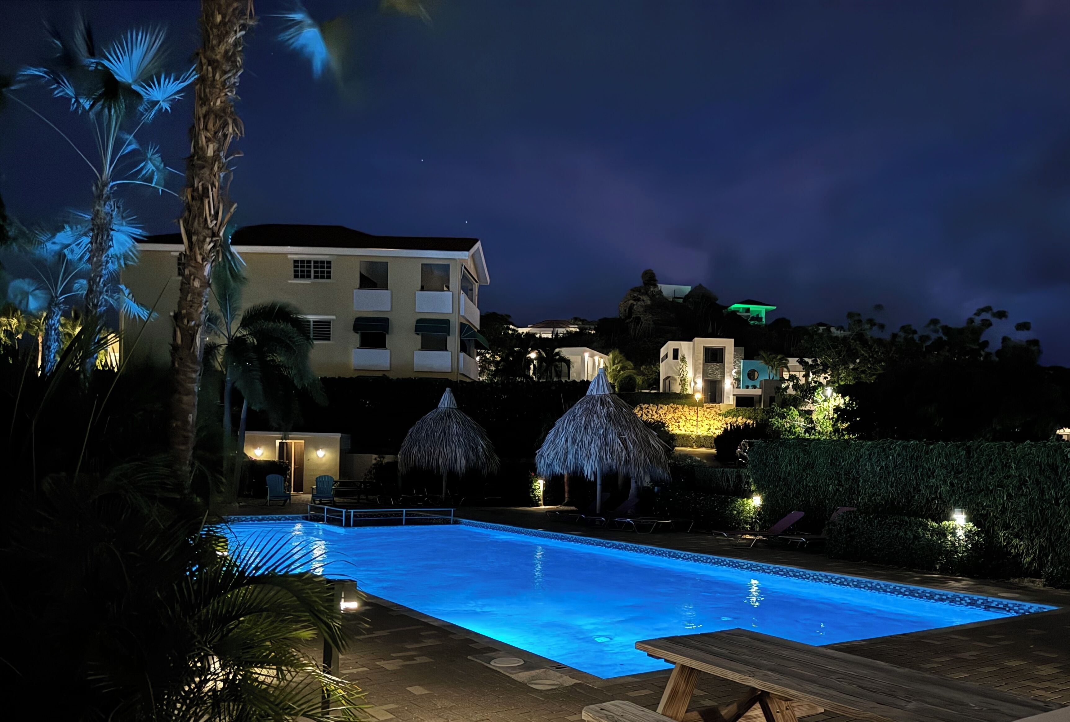 Ein Schwimmbeckenbereich bei Nacht mit klarem Wasser, Liegestühlen und Strohdächern.