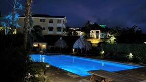 A nighttime swimming pool area with a clear pool, lounge chairs, and thatched umbrellas. - Bluebay - TT#12: Poolside Ocean View - AIRCO  (Willemstad)