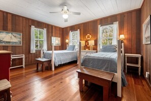 Quarto com duas camas, piso de madeira, ventilador de teto e um quadro na parede.