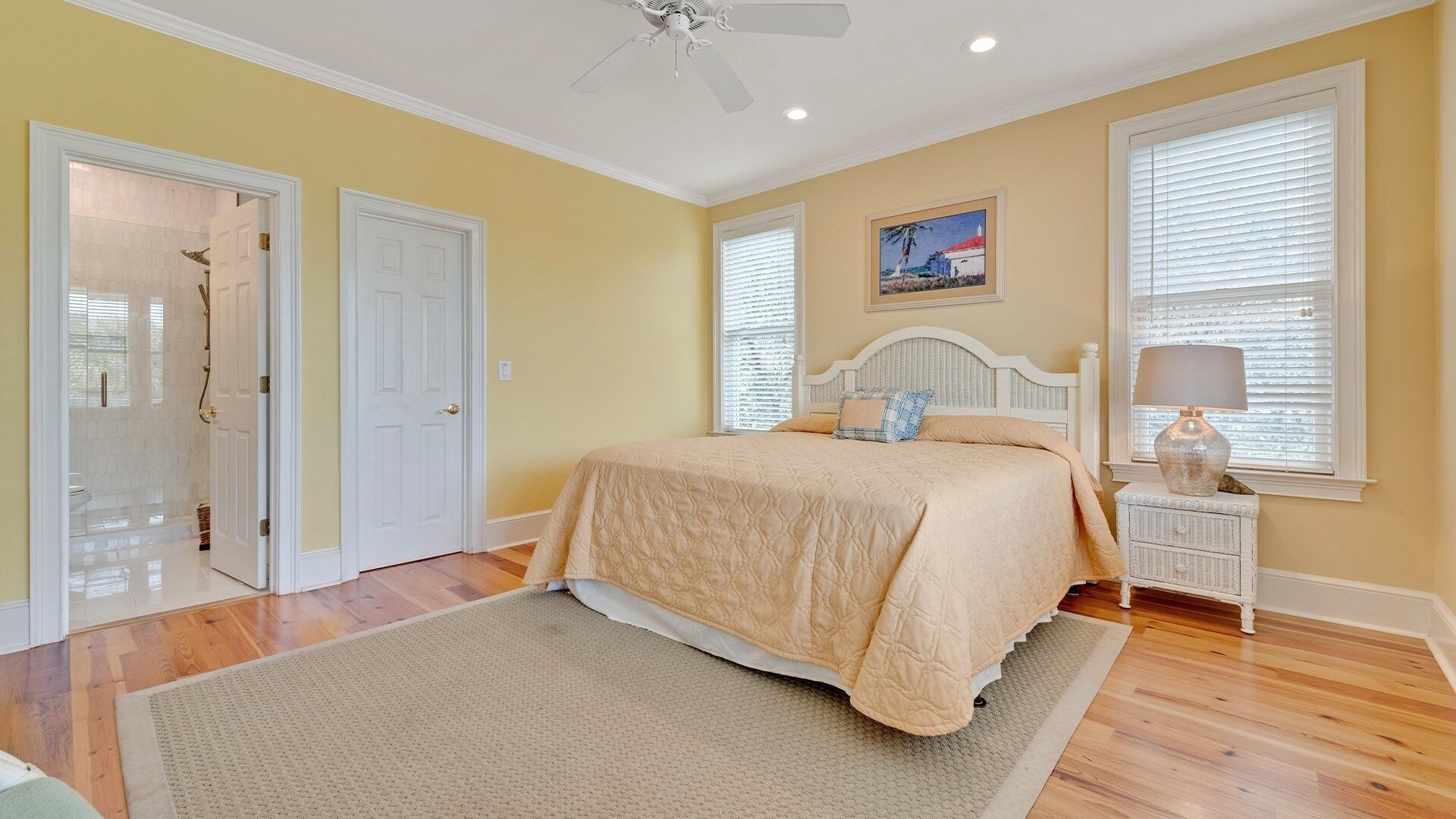 A bedroom with a bed, a nightstand, a lamp, a ceiling fan, and a painting on the wall.