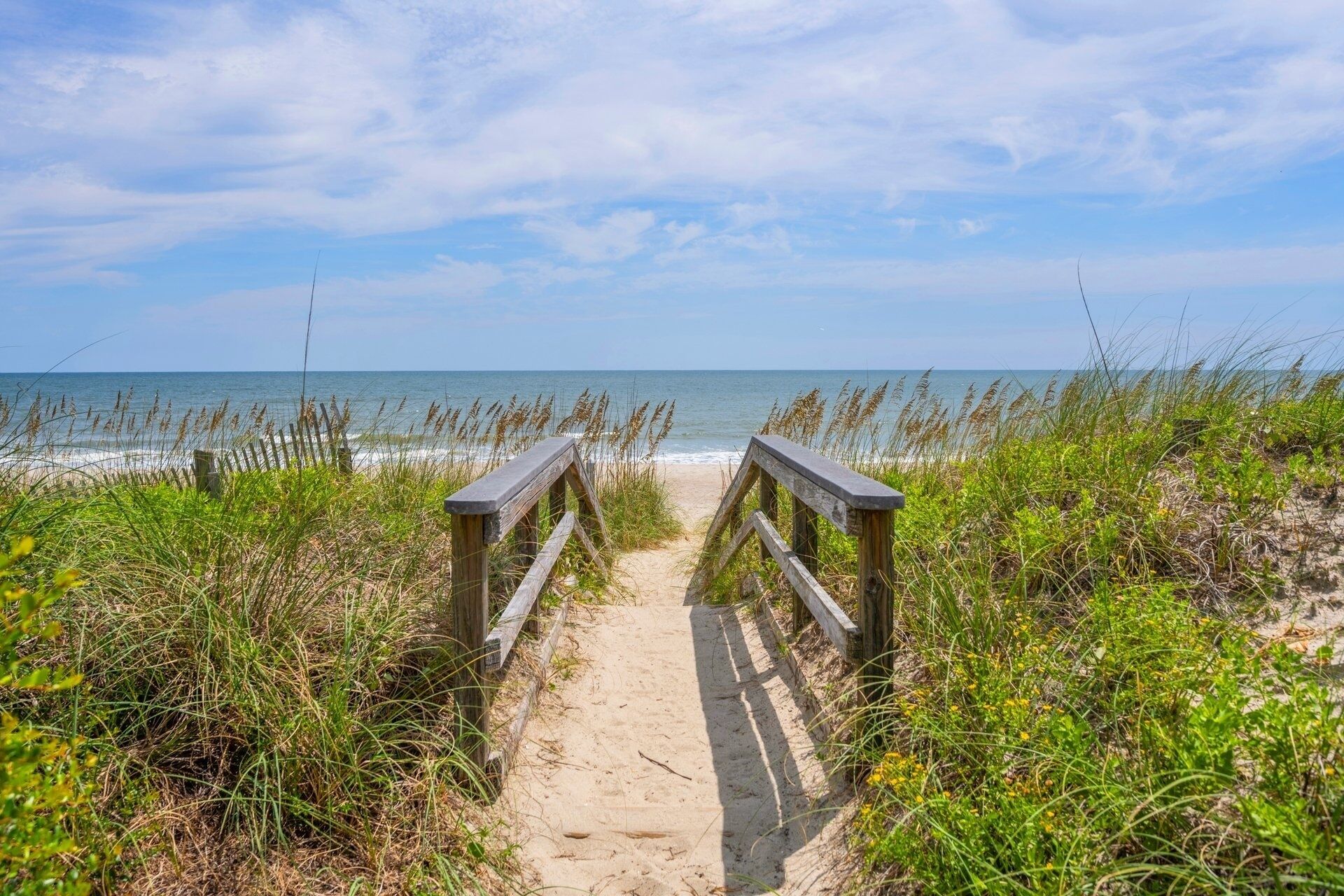 A sandy beach path leading to the ocean, surrounded by tall grasses and wooden railings.