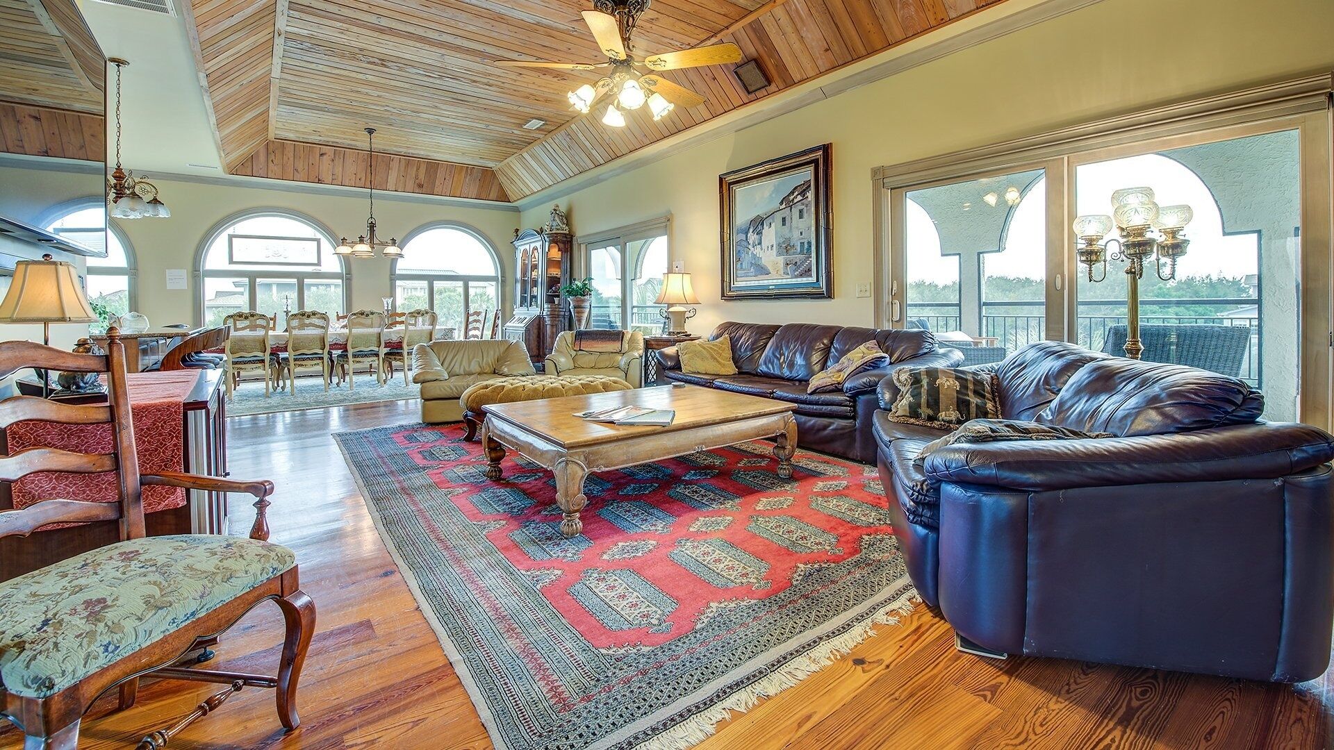 A spacious living room with a high ceiling, wooden floors, leather sofas, a dining area with chairs, and large windows offering views.