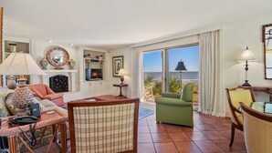 A spacious living room with a large window offering a view of the sea, a fireplace, and a television.