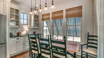 A kitchen with white cabinets, a wooden dining table, and chairs. There are multiple windows with blinds, and a chandelier hangs above the table.