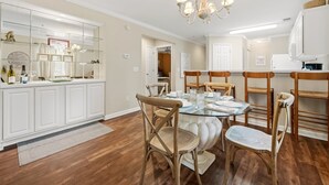 A dining area with a round glass table, wooden chairs, a chandelier, and a kitchen with white cabinets and a glass display case.