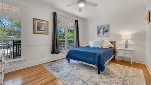 Ein Schlafzimmer mit einem Bett, einem Nachttisch, einer Lampe, einem Deckenventilator und großen Fenstern mit Vorhängen.