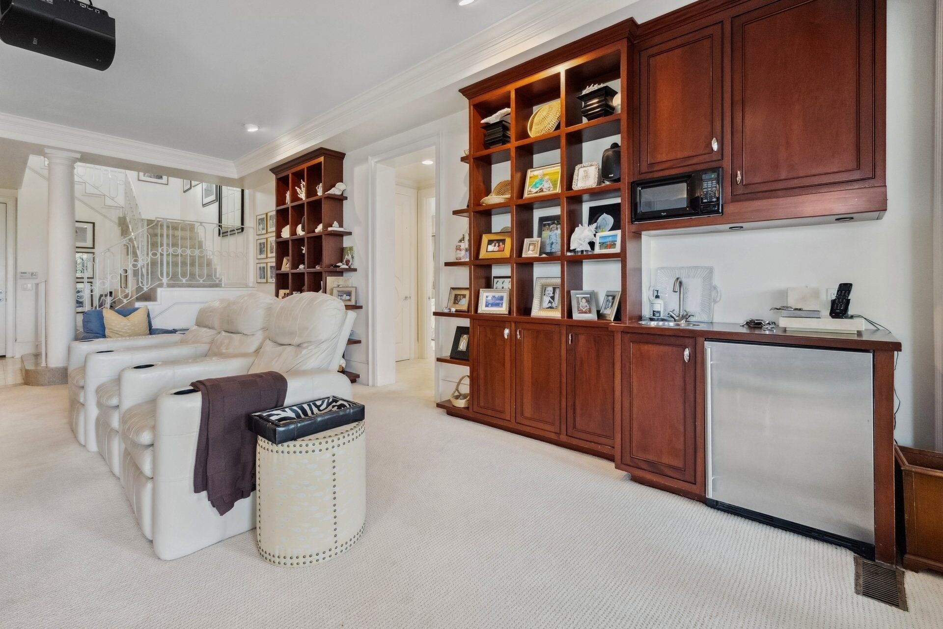 A spacious living room with a white leather sofa, a wooden bookshelf filled with decorative items, a small refrigerator, and a white coffee table.