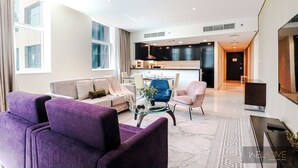 A modern living room with purple sofas, a glass coffee table, and a kitchen in the background.