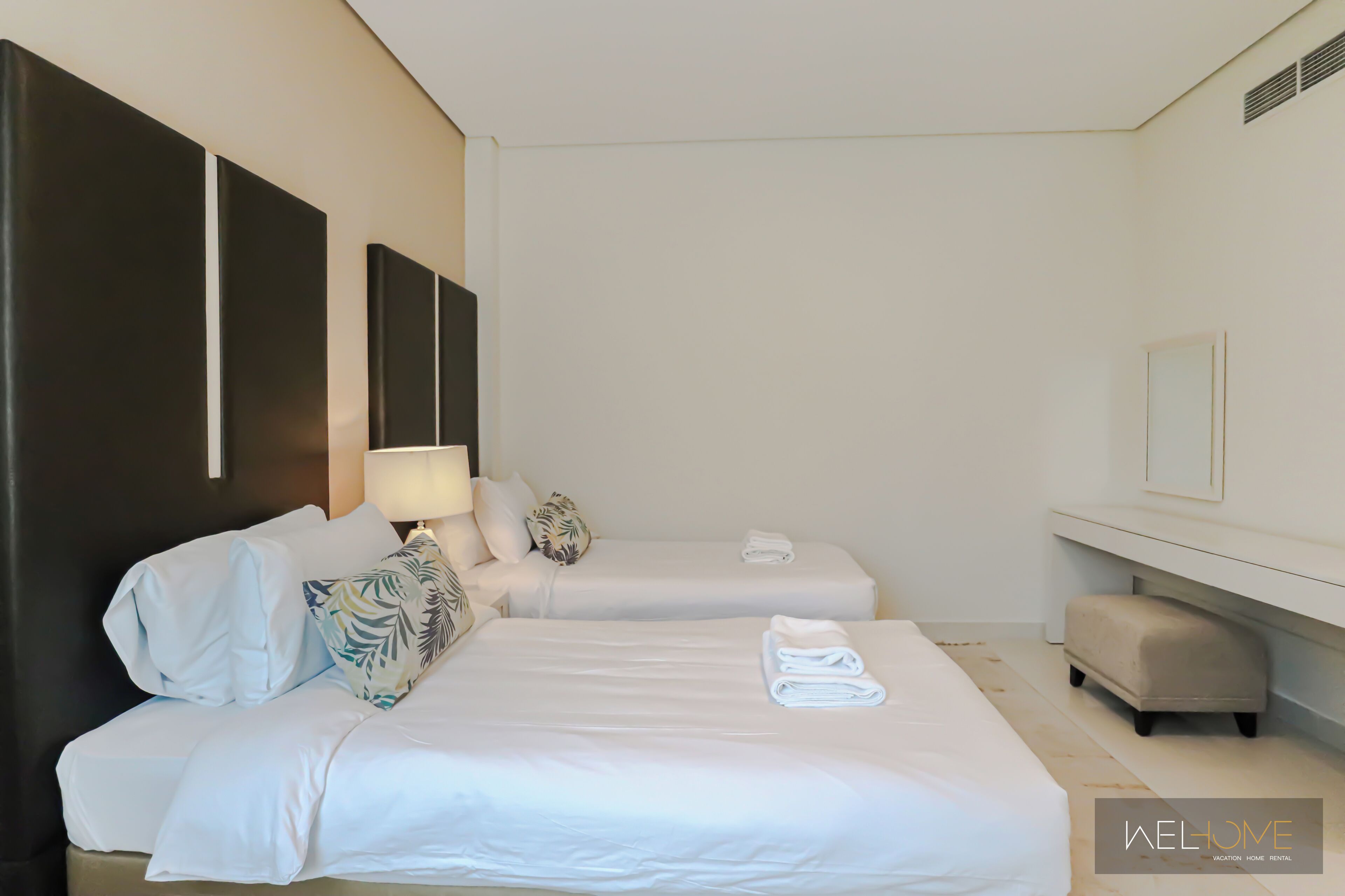A hotel room with two beds, a bedside table with a lamp, a bench, and a mirror on the wall.