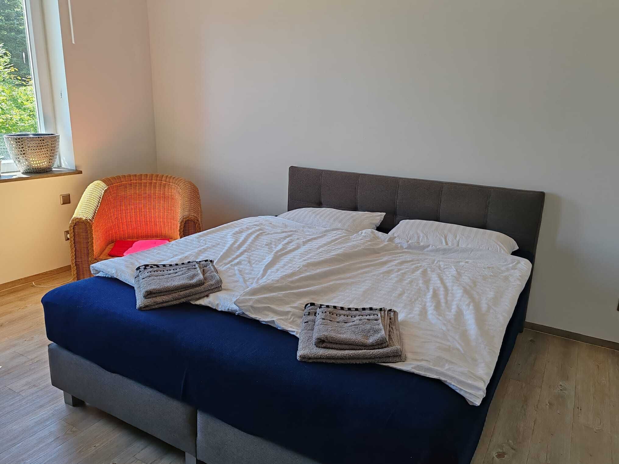 Un letto con lenzuola bianche e due asciugamani piegati, una panca rivestita in grigio e una sedia in vimini con cuscino.