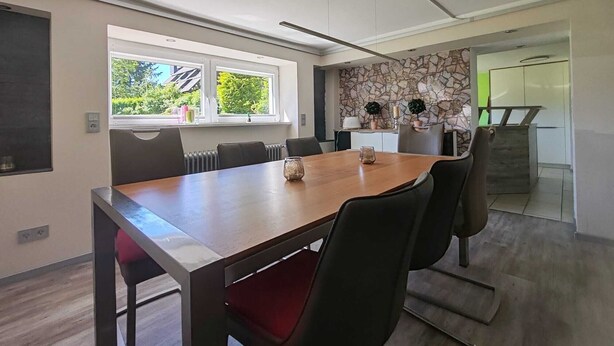 A modern dining area with a wooden table and black chairs, a stone wall, and a view of greenery outside. - "Fuchsbau" vacation apartment for up to 8 people in the Oberharz (Goslar)