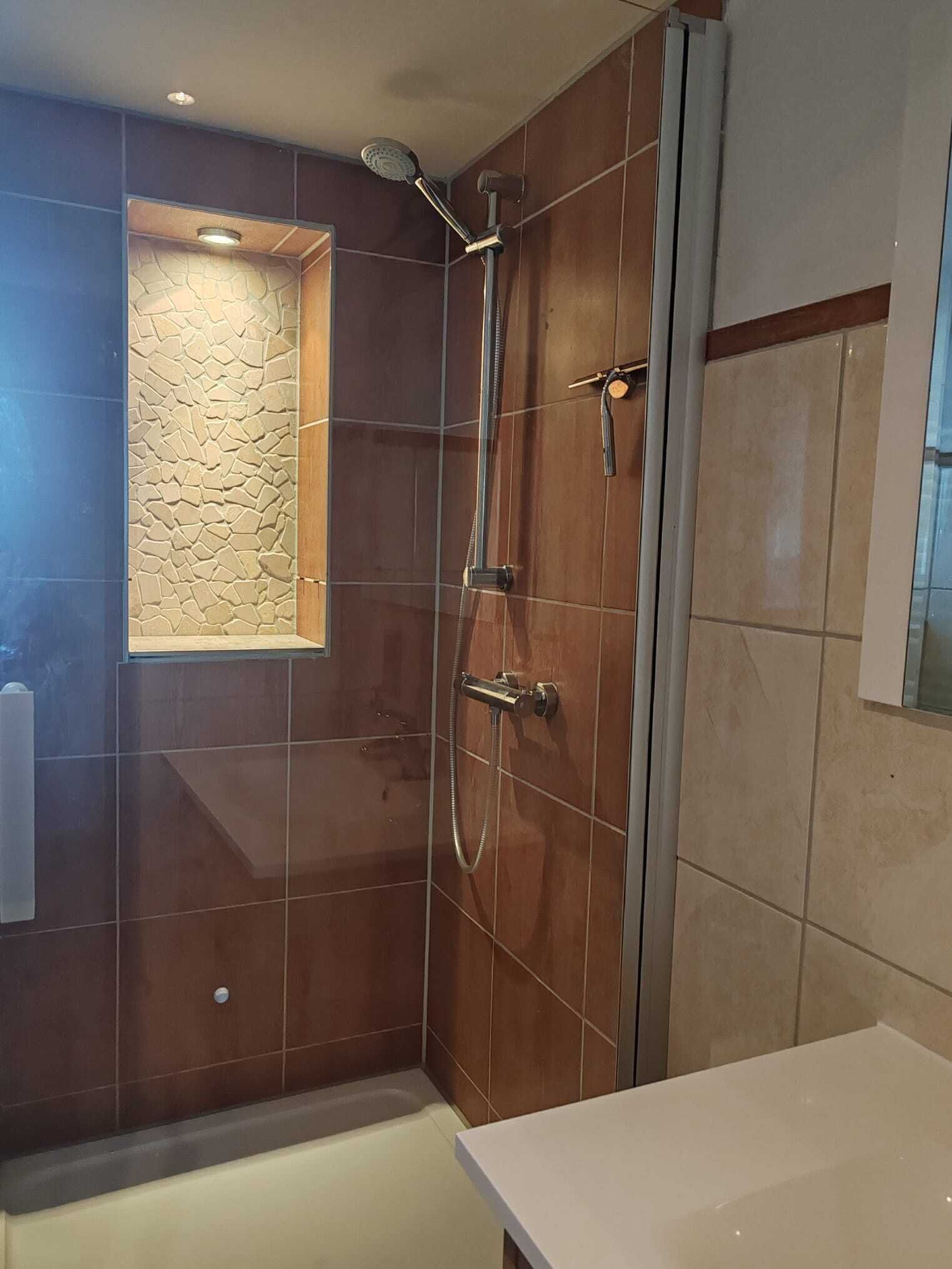 Ein modernes Badezimmer mit einer gläsernen Duschkabine, einem kleinen Fenster mit Steinoptik und einem weißen Waschbecken.