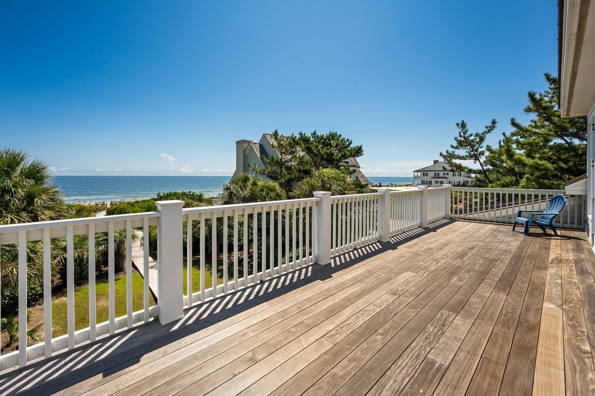 A wooden deck with a white railing, overlooking a beach with ocean and houses in the background.