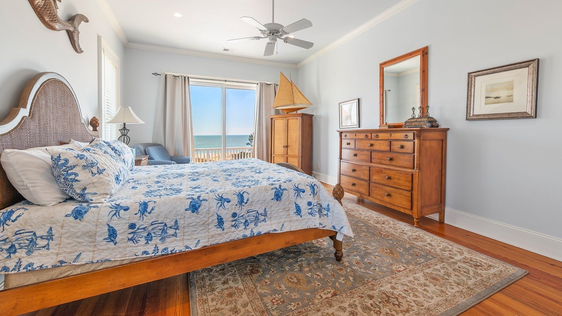 A bedroom with a bed, a dresser, a ceiling fan, a view of the ocean, and a large window.