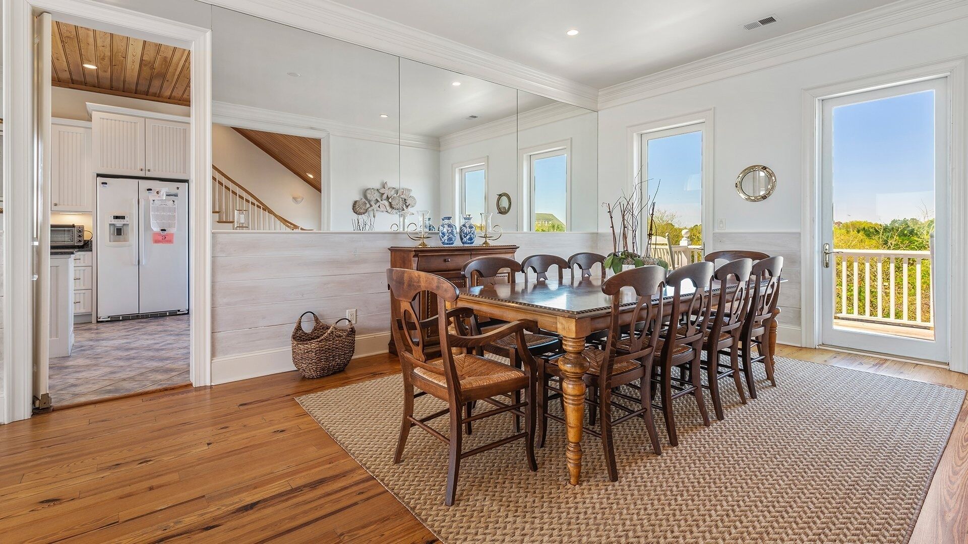 Un vaste espace de salle à manger avec une table et des chaises en bois, une cuisine dotée de meubles blancs et une vue sur l’extérieur grâce à de grandes fenêtres.