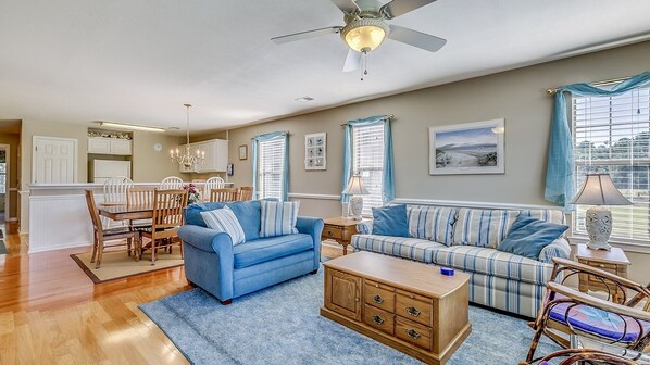 A spacious living room with a blue sofa, a striped sofa, a wooden chest of drawers, and a dining area with wooden chairs and a white table.