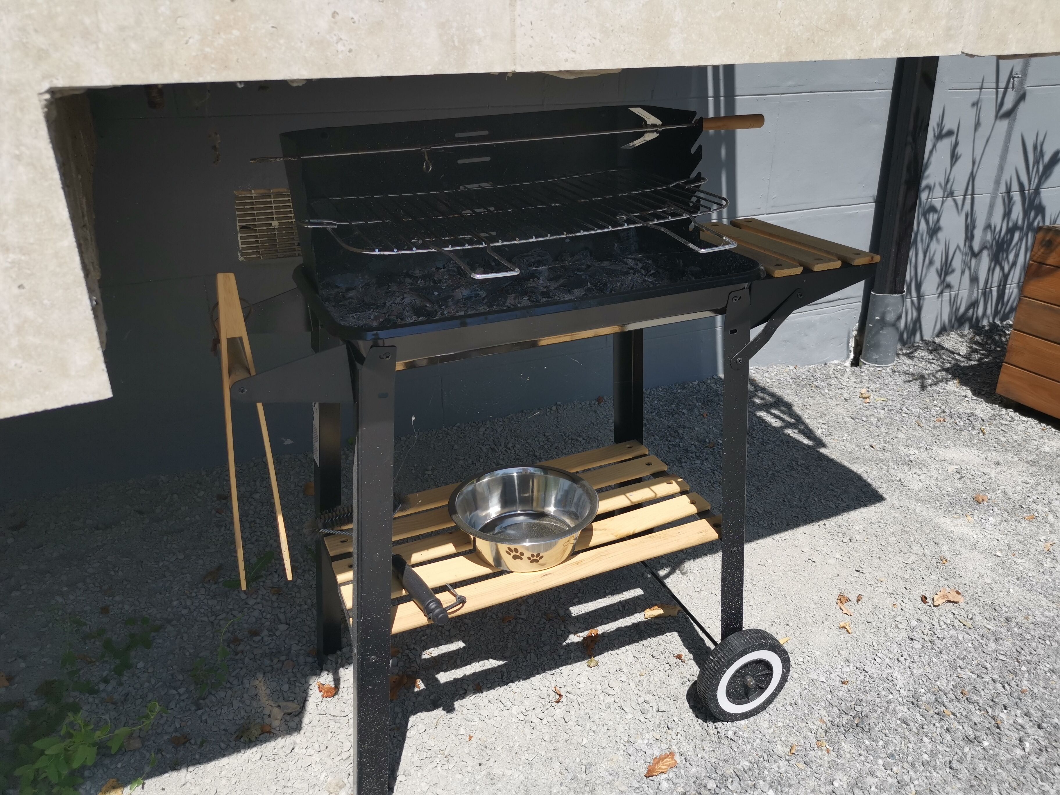 Una parrilla portátil con un recipiente metálico sobre un carro con ruedas, ubicada al aire libre sobre grava.