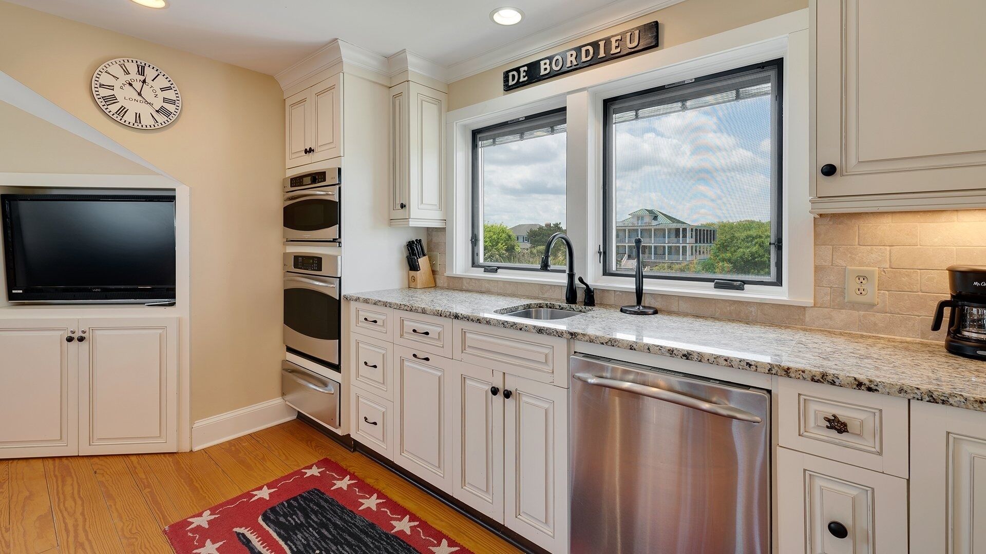 A kitchen with white cabinets, a stainless steel dishwasher, a double sink, and a large window with a view of a house.