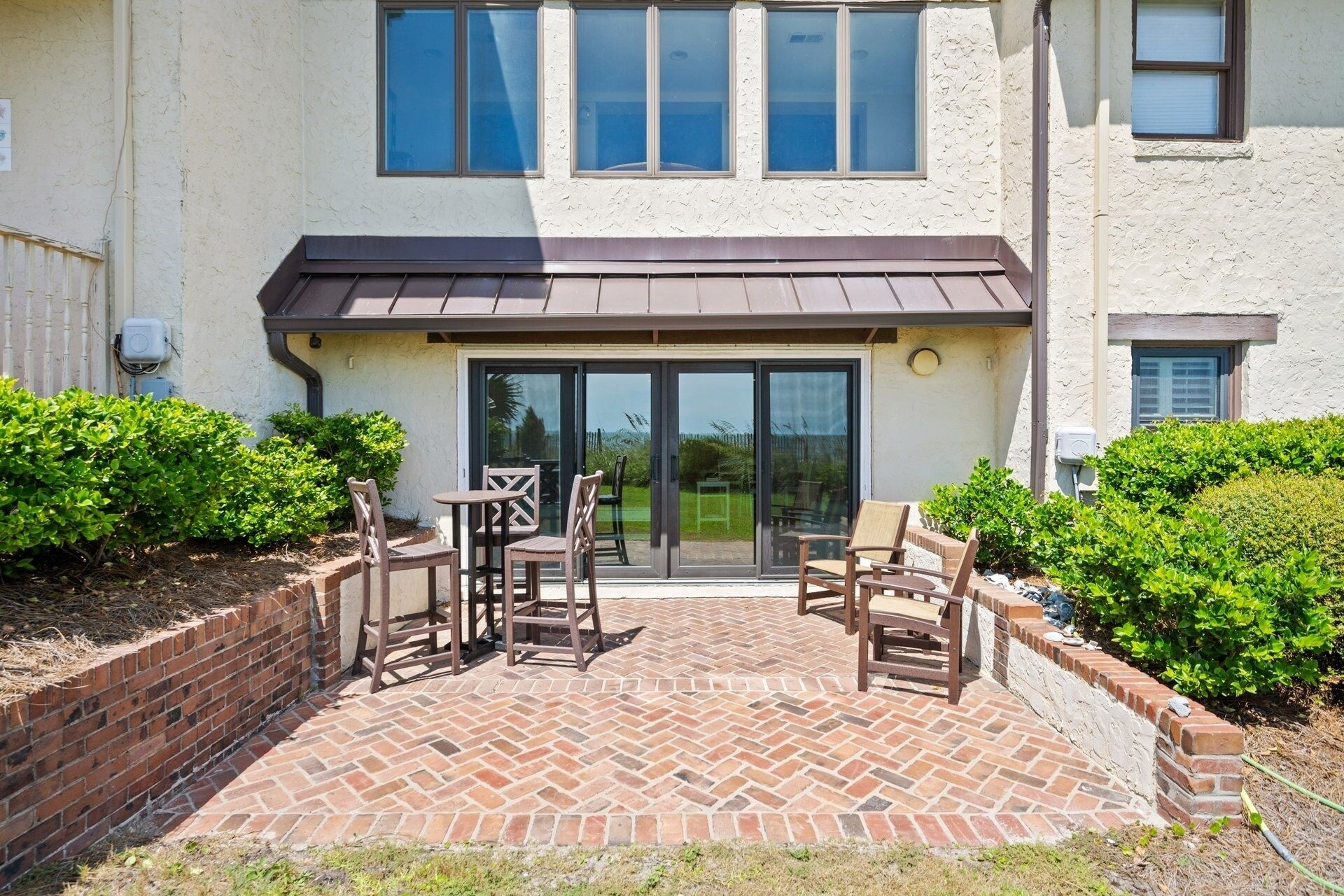 A patio with brick flooring and outdoor furniture, a sliding glass door entrance, and surrounding greenery.