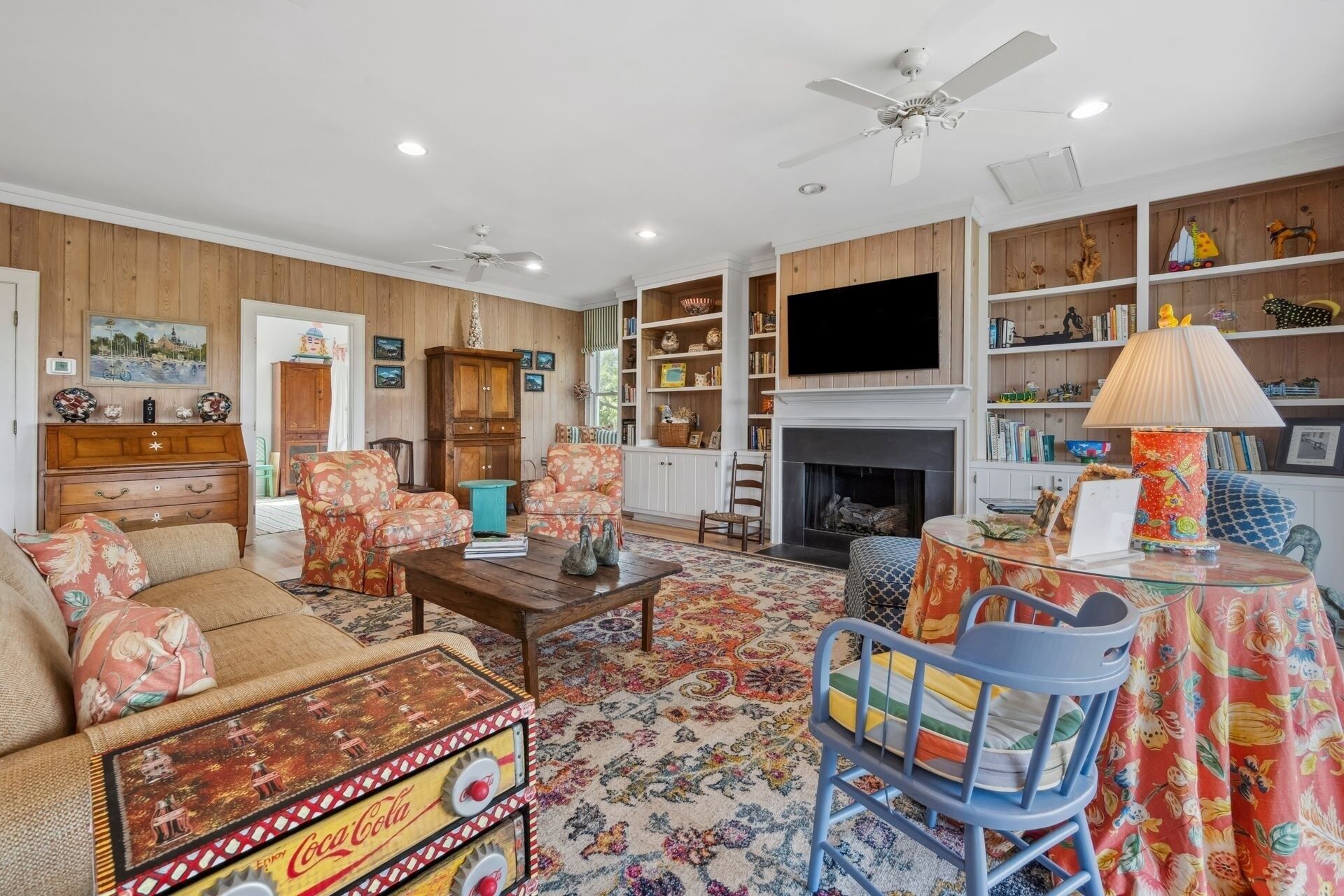 A cozy living room with a fireplace, wooden paneling, and a bookshelf filled with books and decorative items.