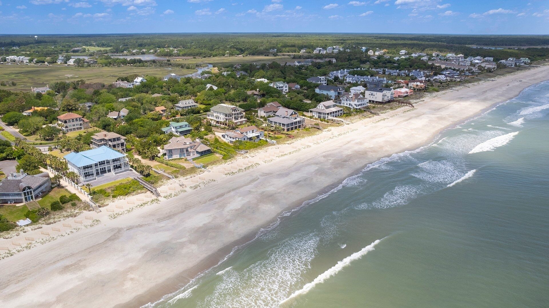 Vue aérienne d’un quartier résidentiel situé sur la côte, avec des maisons le long du bord de mer, une plage de sable et une vue sur l’océan.