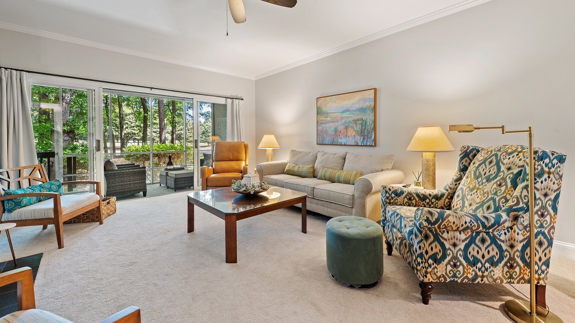 A spacious living room with a large sliding glass door leading to a garden, a sofa set, a patterned armchair, and a painting on the wall.