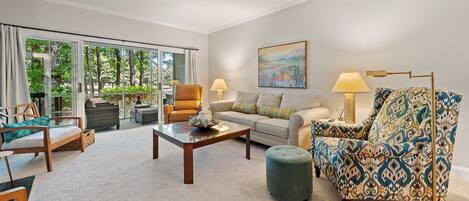 A spacious living room with a large sliding glass door leading to a garden, a sofa set, a patterned armchair, and a painting on the wall.