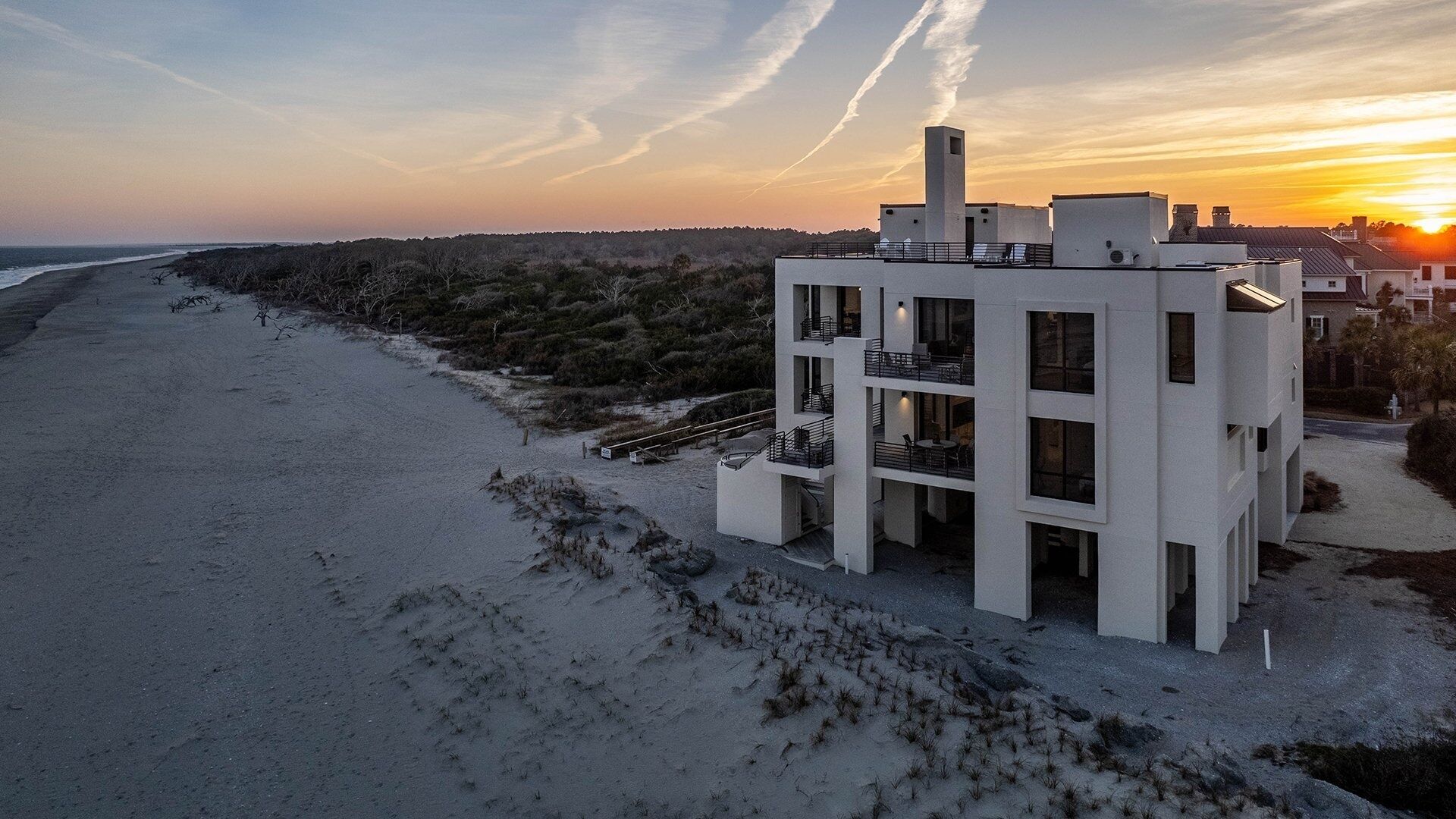 Un edificio moderno de varios pisos con balcones, ubicado en una playa con vista al océano y al atardecer.
