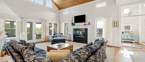 A spacious living room with a high ceiling, large windows, a fireplace, and a television.