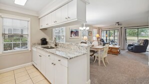 A modern kitchen with white cabinetry, a marble countertop, and a dining area with a table and chairs. - Fore Your Pleasure At Pawleys Plantation 2 Bedroom Condo by Peace Vacations (Pawleys Island)