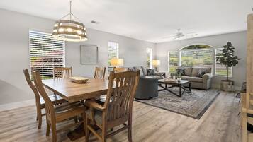 A spacious living room with a dining area, a ceiling fan, and large windows with blinds.