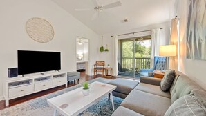 A modern living room with a flat-screen TV, a white coffee table, a grey sofa, and a balcony with a view of greenery. - Welcome To True Blue 52 F 2 Bedroom Condo by Peace Vacations (Pawleys Island)
