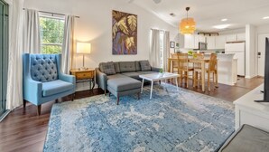 A modern living room with a blue armchair, a grey sofa, a coffee table, and a dining area with wooden chairs and a table. - Welcome To True Blue 52 F 2 Bedroom Condo by Peace Vacations (Pawleys Island)