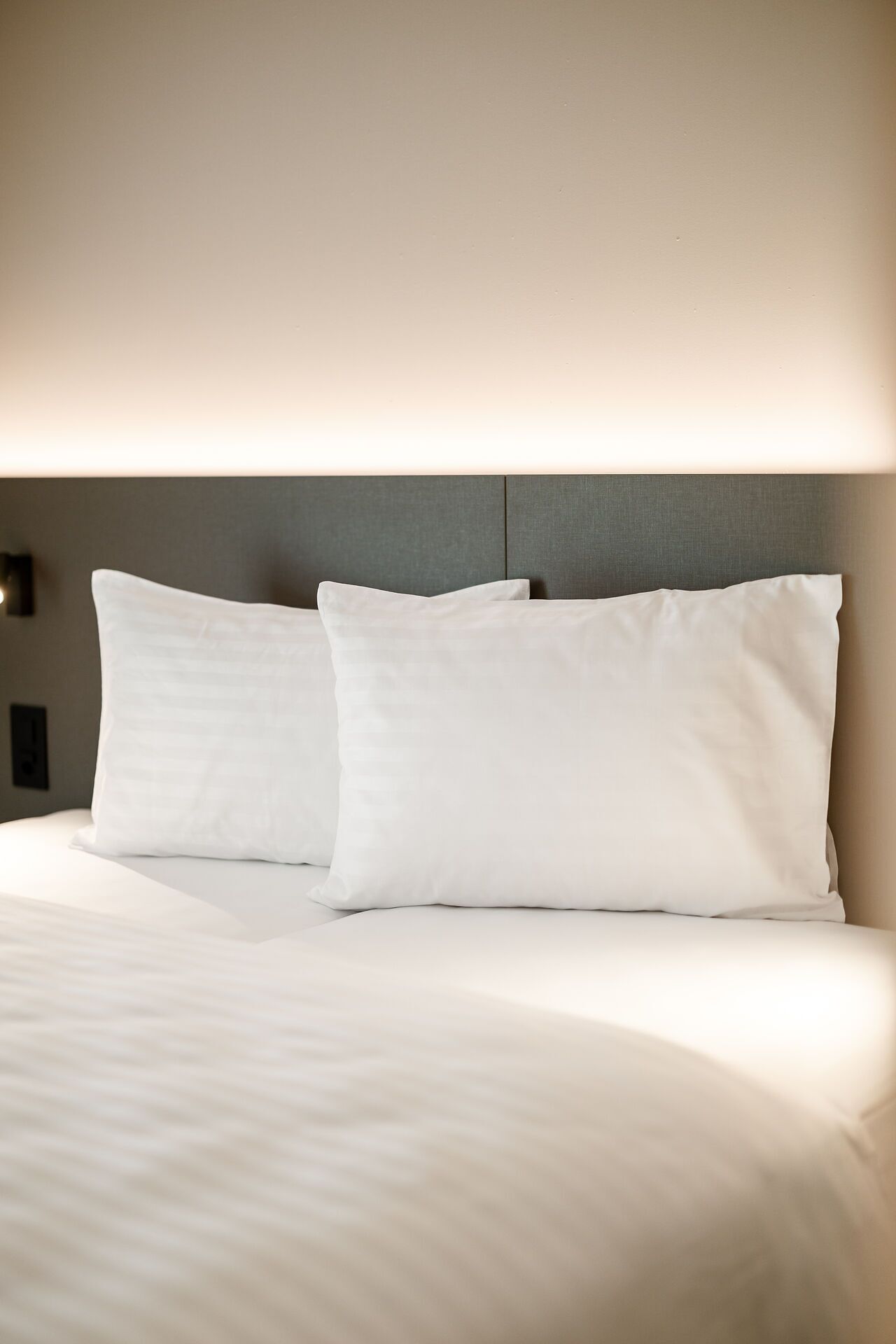 Una cama bien hecha con sábanas blancas y dos almohadas, frente a una pared lisa con una lámpara discreta arriba.