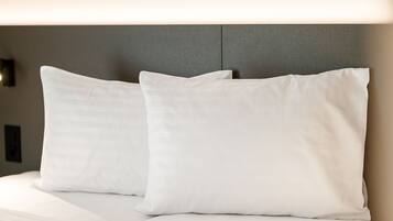 Una cama bien hecha con sábanas blancas y dos almohadas, frente a una pared lisa con una lámpara discreta arriba.