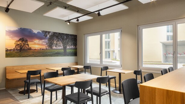 A modern dining area with wooden tables and black chairs, large windows, and a sunset painting on the wall. - bsmart motel Amriswil (Amriswil)