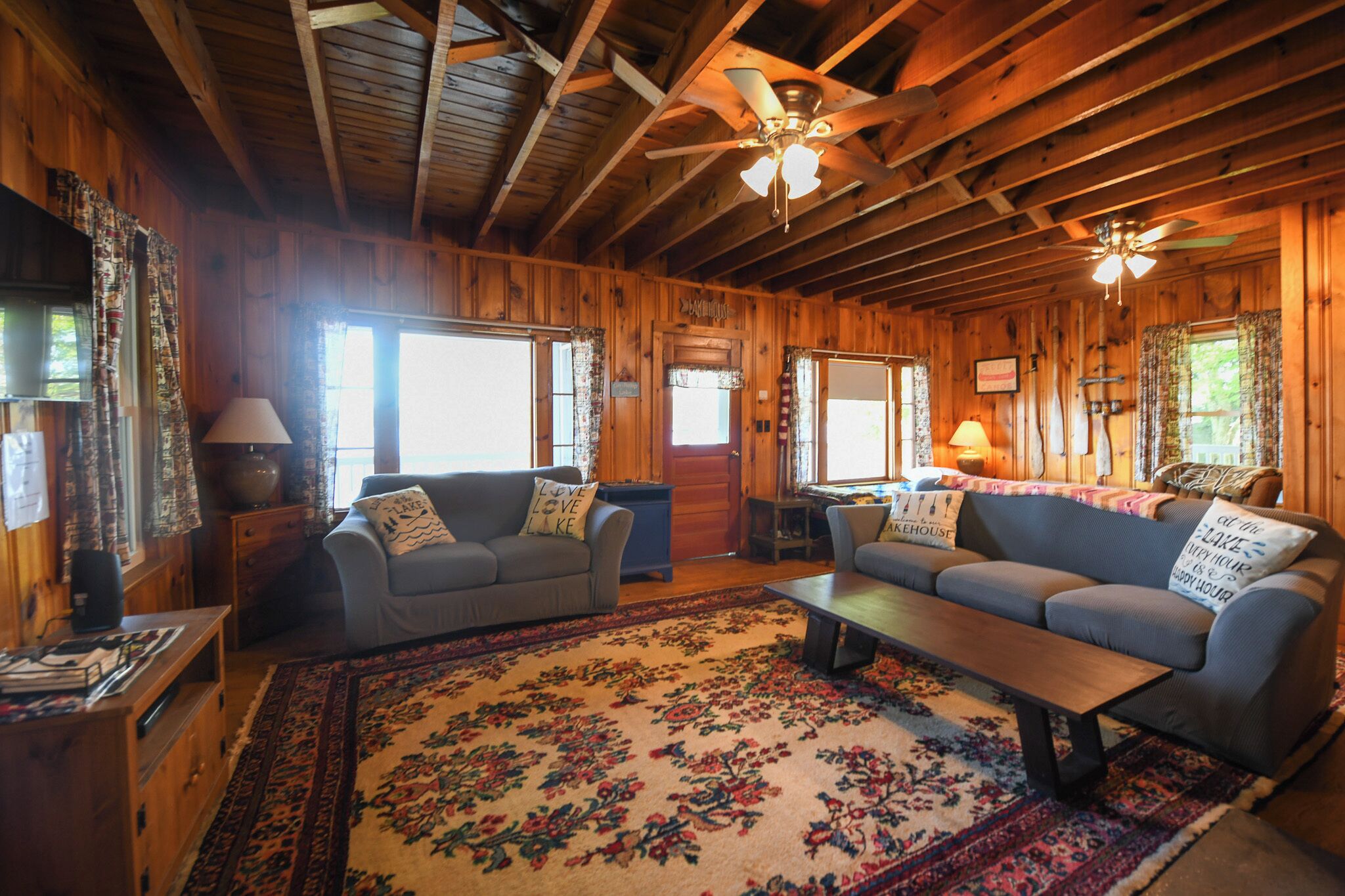 Interior de uma cabana de madeira com um tapete floral grande, dois sofás, uma mesa de centro e um ventilador de teto.