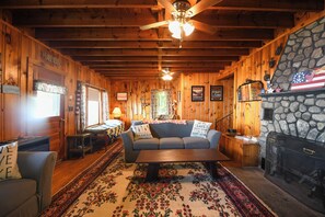 A cozy cabin interior with wooden walls, a stone fireplace, a sofa with pillows, a coffee table, and a ceiling fan.