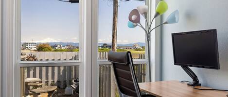 A room with a desk, chair, and a computer monitor. There is a window with a view of a mountain and a fence.