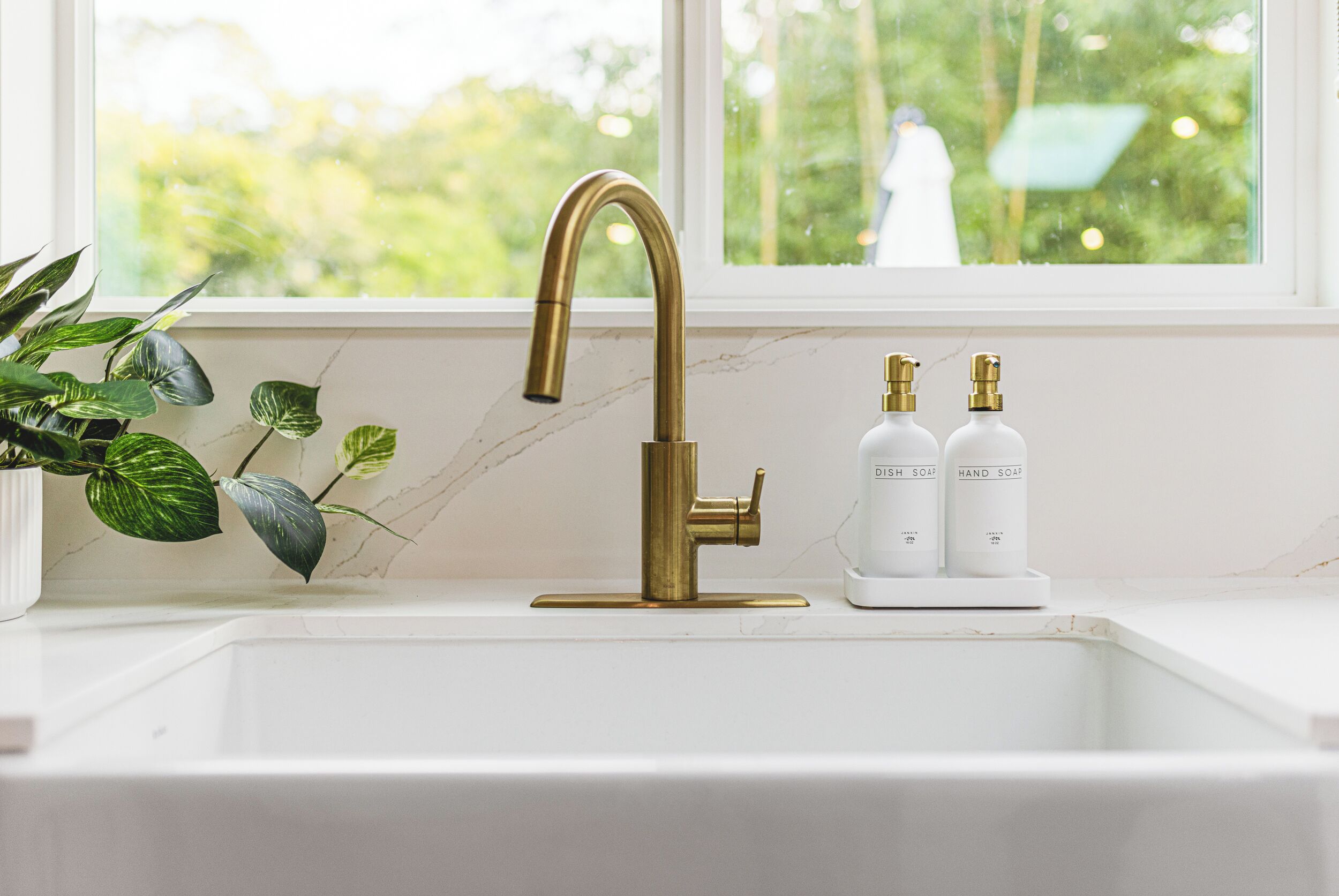A kitchen sink with a gold faucet, two soap bottles, and a potted plant.