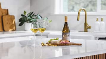 A kitchen counter with a bottle of wine, two glasses, a wooden cutting board with cheese and grapes, and a gold faucet.