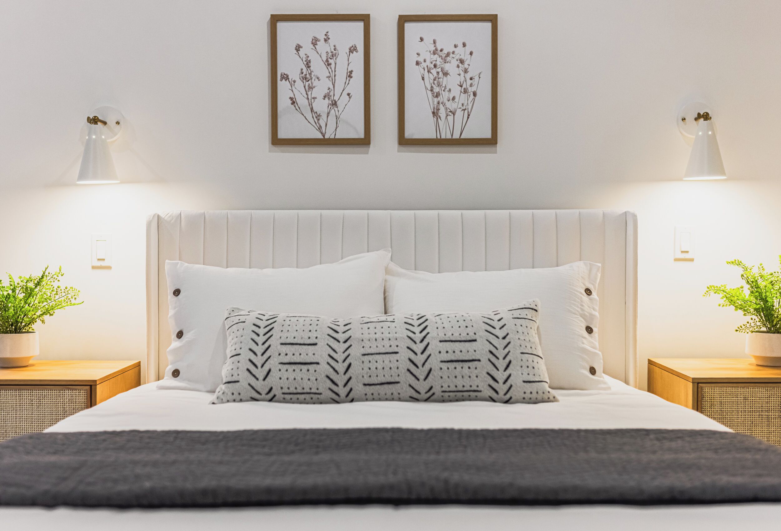 Una cama bien hecha con cabecera blanca, dos cuadros enmarcados y dos lámparas de noche sobre mesitas de madera.