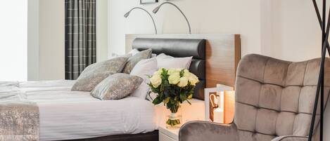 A bedroom with a bed, a bedside table with a lamp, a chair, and a vase of flowers.