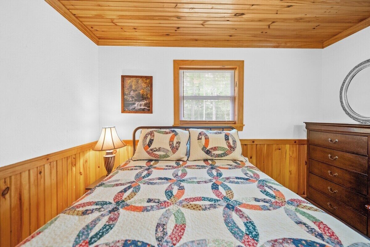 Una camera da letto con soffitto in legno, un letto con copriletto colorato, una finestra con persiane, un comò e uno specchio fissato alla parete.