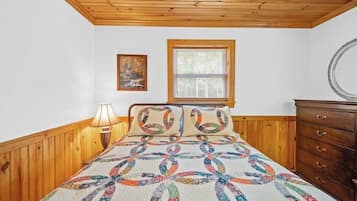 Una camera da letto con soffitto in legno, un letto con copriletto colorato, una finestra con persiane, un comò e uno specchio fissato alla parete.