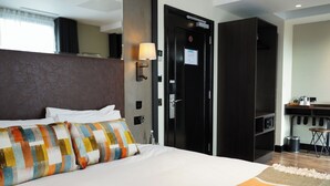 A hotel room with a large bed, a bedside table, a lamp, a wardrobe, and a kitchenette area.