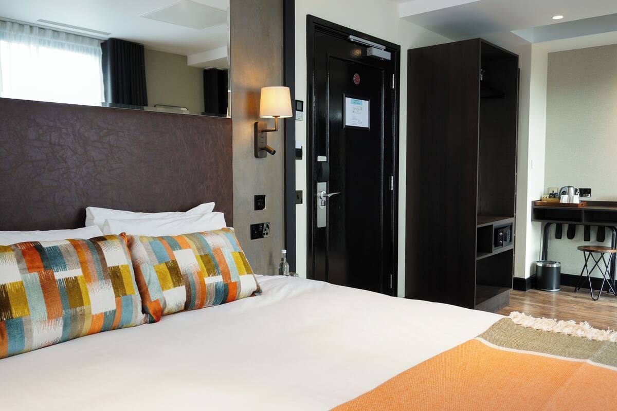 A hotel room with a large bed, a bedside table, a lamp, a wardrobe, and a kitchenette area.
